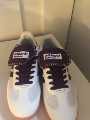 Adidas Wales Bonner vita sneakers - Säljer ett par Adidas Wales Bonner sneakers med vit ovandel, mörklila detaljer och klassiska tre ränder på sidan. Skorna har bruna snören, bred tunga med kontrastsöm och gummisula. Perfekt för dig som vill ha en unik och stilren sneaker.