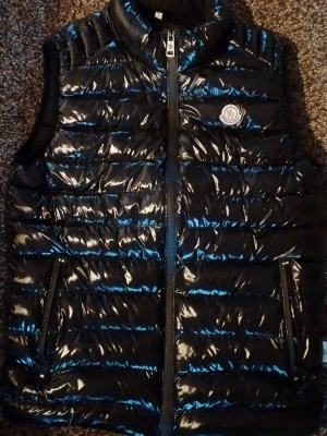 Svart glansig dunväst monclr size S/M - Snygg svart dunväst från Monclr med glansigt material och klassisk logga på bröstet. Västen har hög krage, två dragkedjefickor och quiltad design. Perfekt för lager-på-lager och streetstyle.