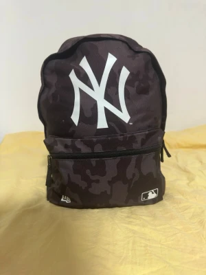 New Era Yankees ryggsäck - Snygg svart ryggsäck från New Era med stort vitt New York Yankees-tryck på framsidan. Väskan har mörkt camouflagemönster, justerbara axelband och en stor framficka med dragkedja. Perfekt för skolan eller stan.