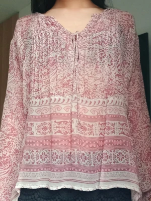 Bohemisk mönstrad blus i rosa toner - Luftig blus med bohemiskt mönster i rosa och vitt. Blusen har v-ringning med knytband och fina rynkade detaljer upptill. Långa, vida ärmar och lätt transparent material ger en avslappnad och trendig look.
