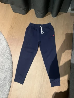 Mörkblå mjukisbyxor från Polo Ralph Lauren - Snygga mörkblå mjukisbyxor från Polo Ralph Lauren med vit dragsko i midjan och röd logga broderad på vänster sida. Byxorna har muddar vid bensluten och är tillverkade i mjukt bomullsmaterial för maximal komfort.