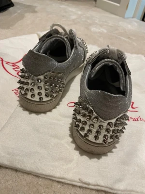 Glittriga Louboutin sneakers med nitar - Snygga sneakers från Christian Louboutin i silverfärgat glittrigt material med coola silvriga nitar över hela hälen. Skorna har lågt skaft och vita snören. Perfekta för dig som vill sticka ut och addera lite edge till din outfit.