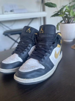 Nike Air Jordan 1 svart/vit/guld - Säljer ett par Nike Air Jordan 1 sneakers i svart, vitt och guld. Skorna har högt skaft, svart läderdetaljer, vit tåbox och sidor samt guldfärgade swoosh och loggor. Klassisk snörning och ikonisk Jordan-design. Perfekta för dig som gillar streetwear. Priset är fast!