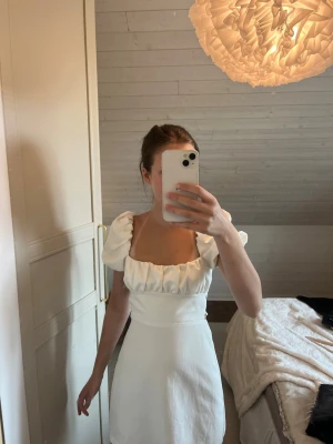 Vit kort klänning från zara - Supersöt vit kort klänning med puffiga kortärmar, fyrkantig urringning och öppen rygg med fin snörning från zara. Klänningen har en figurnära passform i midjan och är gjord i ett mjukt bomullsliknande material. Perfekt för sommarens alla tillfällen! Jag har endast använt den en gång och är i mycket fint skick.