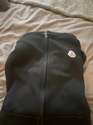 Svart zip hoodie från Moncler - Säljer en svart hoodie från Moncler med dragkedja framtill och klassisk Moncler-logga på bröstet. Hoodien har huva med dragsko och fickor framtill. Perfekt för dig som gillar stilrena och exklusiva plagg.