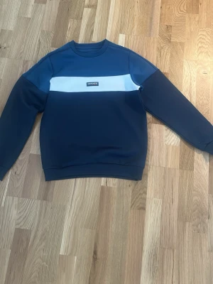 Blå och vit stickad tröja McKenzie - Snygg blå stickad tröja från McKenzie med vit panel över bröstet och logga framtill. Tröjan har rund halsringning och långa ärmar. Perfekt för en avslappnad och stilren look. Storlek L i barn, passar ungefär XS i herr