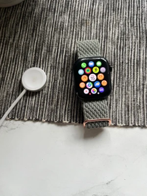 Apple Watch  - Apple Watch Nike med svart fyrkantig urtavla och grått flätat textilarmband. Klockan har en stor, tydlig display och kommer med laddare. Perfekt för dig som gillar smarta funktioner och sportig design.