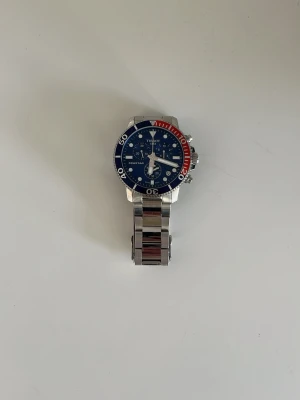 Tissot blå och röd klocka i stål - Snygg Tissot klocka med blå urtavla och detaljer i rött och vitt. Klockan har en robust länk i rostfritt stål och tydliga visare. Perfekt accessoar för dig som gillar sportig och stilren design. Klockan är en 45mm och urverket är quartz (batteridrivet). Inköpt hos stjärnurmakaren för ca ett år sedan (kvitto finns).