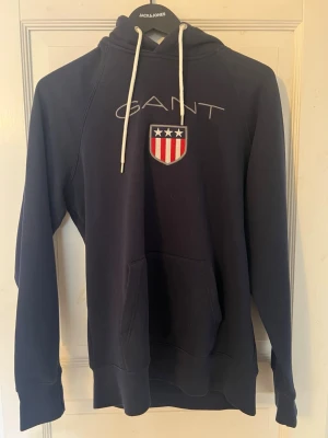 Mörkblå hoodie från GANT med logga - Snygg mörkblå hoodie från GANT med vit dragsko i huvan och stor broderad logga samt sköld med stjärnor och ränder på bröstet. Klassisk känguruficka framtill och ribbade muddar vid ärmslut och nederkant. Perfekt för en avslappnad och sportig stil.