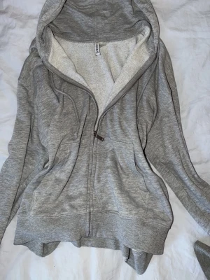 Grå zip hoodie med dragsko - Mysig grå hoodie från Fruit of the Loom med dragkedja framtill, två fickor och justerbar huva med dragsko. Insidan är mjukt fodrad i ljust material och muddar finns vid ärmslut och nederkant. Perfekt för chill dagar eller när du vill vara extra bekväm.