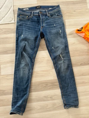 Blå slitna slim jeans Jack & Jones - Snygga blå slim fit jeans från Jack & Jones, modell Glenn. Jeansen har slitningar och en tvättad look med klassiska fem fickor och knappgylf. Perfekta för en avslappnad och trendig stil.