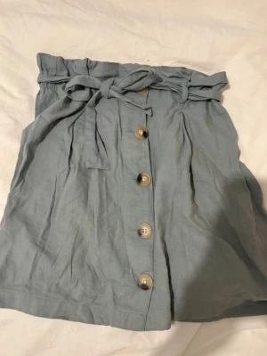 Ljusblå kortkjol med knappar Gina Tricot - Supersöt ljusgrön kortkjol från Gina Tricot med knyt i midjan och dekorativa knappar framtill. Kjolen har en papperspåse-midja och är gjord i linne, lätt material som passar perfekt till varmare dagar.