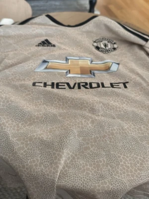 Manchester United bortatröja Adidas - Snygg beige Manchester United bortatröja från Adidas med svart logga och detaljer. Tröjan har ett unikt mönster, klubbmärke på bröstet och Chevrolet som sponsortryck. Perfekt för dig som älskar fotboll och vill sticka ut på planen.