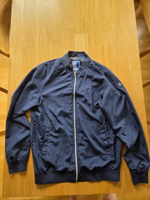 Jack & Jones marinblå jacka , storlek 176 - Jacka från Jack & Jones med dragkedja framtill. Jackan har en rundad krage och muddar vid ärmslut och nederkant. Den har två fickor fram med tryckknappar. Insidan är fodrad med ett mönstrat tyg i mesh.