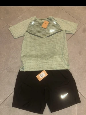 Nike träningsset t-shirt och shorts - Snyggt träningsset från Nike med en ljusgrön, ventilerad t-shirt i meshmaterial och svarta shorts. Båda plaggen har reflekterande Nike-logga och är tillverkade i lätt, snabbtorkande material som passar perfekt för löpning eller gym.