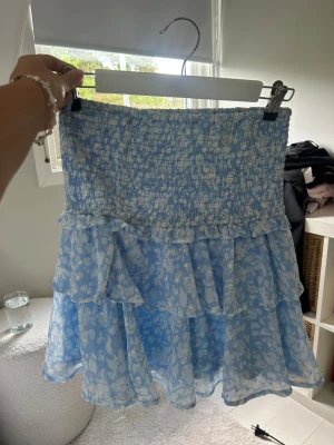 Blå volangkjol med blommönster - Supersöt blå kjol med vita blommor och volanger. Kjolen har smock i midjan för skön passform och dubbla lager volanger som ger en luftig och lekfull look. Perfekt för dig som gillar romantisk och trendig stil.