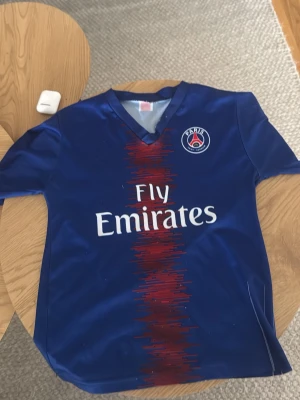 PSG fotbollströja Mbappé #7 blå - Snygg blå PSG matchtröja med Mbappé och nummer 7 på ryggen. Tröjan har klubbmärke på bröstet, Fly Emirates-tryck och röd detalj i mitten. Tillverkad i lätt och ventilerande material, perfekt för fotboll eller träning.