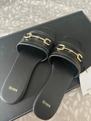 Svarta sandaler med gulddetaljer från Mango - Stilrena svarta sandaler från Mango med två breda remmar i konstläder och snygga guldfärgade metallspännen. Sandalerna har fyrkantig tå och vita kontrastsömmar som ger en modern touch. Perfekta för en trendig och avslappnad look.