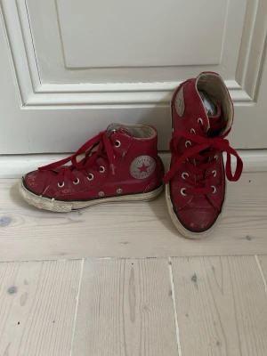 Röda Converse Chuck Taylor Highs - Klassiska röda Converse Chuck Taylor All Star high tops med vita sulor och röda skosnören. Skorna har den ikoniska stjärnloggan på sidan och metallfärgade öljetter. Tillverkade i canvas med gummisula och svart rand runt sulan. Perfekta för en avslappnad streetstyle.