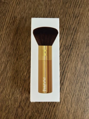 BeautyAct puderborste  - Lyxig puderborste från BeautyAct by KICKS i snygg guldfärg med täta, mjuka syntetiska borst. Perfekt för att applicera puder jämnt över ansiktet. Borsten har ett stilrent och modernt utseende med rundad form och kompakt handtag.
