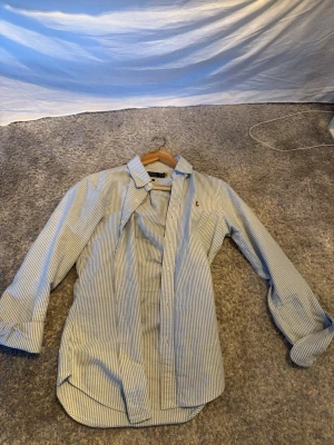 Randig skjorta från Polo Ralph Lauren XS - Klassisk blåvit randig skjorta från Polo Ralph Lauren i slim fit-modell. Skjortan har långa ärmar, knappar framtill och en liten broderad logga på bröstet. Tillverkad i mjuk bomull som känns skön mot huden.