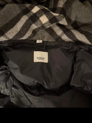 Burberry Jacka  - Snygg svart pufferjacka från Burberry med klassiskt rutigt foder i huvan. Jackan har dragkedja och knappar framtill, två fickor med dragkedja och en stor huva med justerbar dragsko. Perfekt för kalla vinterdagar.