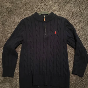 Mörkblå stickad tröja Polo Ralph Lauren - Kabelstickad mörkblå tröja från Polo Ralph Lauren med halv dragkedja och röd broderad logga på bröstet. Tröjan har hög krage, långa ärmar och ribbade muddar. Perfekt för dig som gillar klassisk och stilren look. Den är i bra skick!!🤩kan gå ner vid snabb affär😉