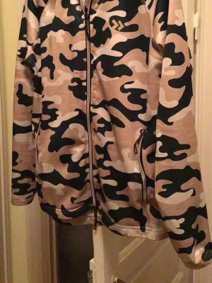 Beige camo vindjacka från 157 - Snygg vindjacka i beige, svart och vit camomönster från 157. Jackan har dragkedja framtill, två sidofickor med dragkedja och mjukt fleecefoder på insidan. Perfekt för dig som gillar streetwear och vill sticka ut med ett coolt mönster. ( några svarta fläckar lite synliga bland mönster)