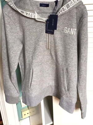 Grå hoodie från GANT med dragkedja - Snygg grå hoodie från GANT med halv dragkedja framtill och vit logga på bröstet. Huva med tryckt text längs kanten och stor känguruficka. Tillverkad i mjukt sweatshirtmaterial, perfekt för en avslappnad look. Strl 170.