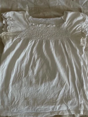 Vit blus med smock och volangärm - Supersöt vit blus med smock upptill och volangdetaljer på de korta ärmarna. Blusen har en luftig passform och är gjord i mjuk bomull, perfekt för en fräsch och enkel look.