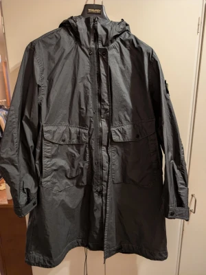 Lång regnjacka från Stone Island - Säljer en mörkgrön parkas från Stone Island med ikonisk patch på ärmen. Jackan har huva, stora fickor framtill och dragsko i nederkant. Tillverkad i lätt, slitstarkt material som ger en clean och modern look. Perfekt för dig som gillar streetwear och funktionella plagg.
