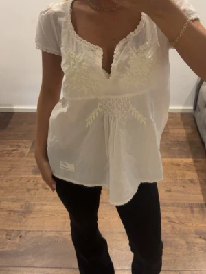 Ljusrosa broderad blus med spetskant - Söt ljusrosa blus med korta ärmar och v-ringning, dekorerad med broderade blommor och spetskant runt halsen. Blusen har smockdetalj under bysten och är tillverkad i ett lätt, luftigt tyg som ger en romantisk känsla.