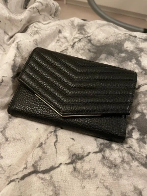 Svart plånbok  - Svart plånbok från Ted Baker i präglat läder med gulddetaljer. Plånboken har ett kuvertformat lock med stilrena sömmar i chevronmönster och diskret logga i guld. Har använts en del men plånboken är ändå i väldigt bra skick.