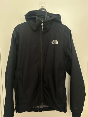 Svart regnjacka från The North Face - Snygg svart regnjacka från The North Face med huva och dragkedja framtill. Jackan har meshfoder på insidan och logga på bröstet. Tillverkad i lätt och vattenavvisande material, perfekt för regniga dagar. Modellen är enkel och stilren med justerbar nederkant.