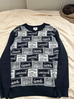 Mönstrad stickad tröja Lacoste Live - Säljer en svart och vit stickad tröja från Lacoste Live med grafiskt mönster och logotyper över hela framsidan. Tröjan har rund halsringning, långa ärmar och ribbade muddar. Perfekt för dig som gillar streetwear och vill sticka ut.