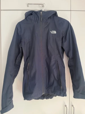 Mörkblå regnjacka från The North Face - Snygg mörkblå regnjacka från The North Face med luva och hel dragkedja framtill. Två praktiska fickor med dragkedja och justerbar nederkant. Jackan är tillverkad i ett vattenavvisande material och har logga på bröstet och ryggen. Perfekt för regniga dagar.