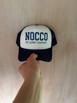NOCCO truckerkeps i blå och vit - Säljer en snygg truckerkeps från NOCCO i blått och vitt med stort tryck framtill. Kepsen har mesh på sidorna och justerbar snapback baktill. Perfekt för dig som gillar sportig stil och vill sticka ut med ett tydligt varumärke.
