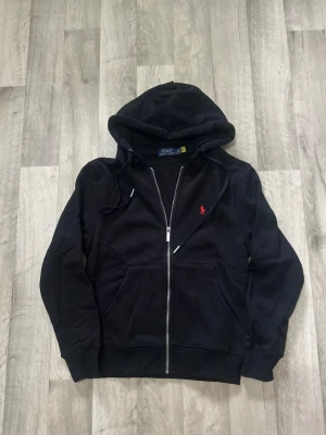 Svart hoodie från Polo Ralph Lauren - Klassisk svart hoodie med dragkedja från Polo Ralph Lauren. Har huva med dragsko, två fickor framtill och den ikoniska röda loggan broderad på bröstet. Tillverkad i mjukt bomullsmaterial för en skön och avslappnad look.