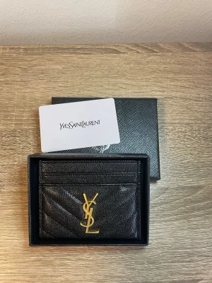 Svart kortfodral från Yves Saint Laurent - Snyggt svart kortfodral från Yves Saint Laurent i präglat läder med guldfärgad YSL-logga framtill. Fodralet har flera kortfack och en stilren, kompakt design. Perfekt för dig som vill ha en lyxig accessoar i fickan.
