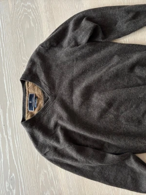 Mörkgrå ulltröja från Hansen & Jacob - Snygg mörkgrå stickad tröja från Hansen & Jacob i 100% lammull. Tröjan har V-ringning och ribbstickade muddar vid hals, ärmslut och nederkant. Perfekt för dig som gillar klassisk och stilren design. Använd ett fåtal gånger och skicket är väldigt bra.