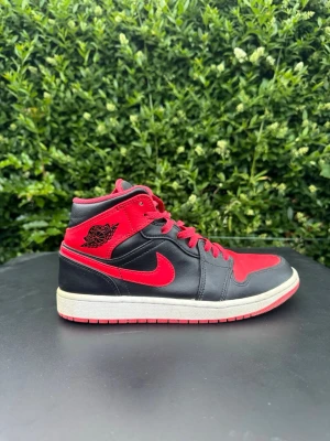 Nike Air Jordan 1 Mid Svart/Röd - Säljer ett par Nike Air Jordan 1 Mid sneakers i svart och rött läder med klassisk röd swoosh och Air Jordan-logga på sidan. Skorna har högt skaft, röda skosnören och vit mellansula. Perfekta för dig som gillar streetwear och ikoniska sneakers.