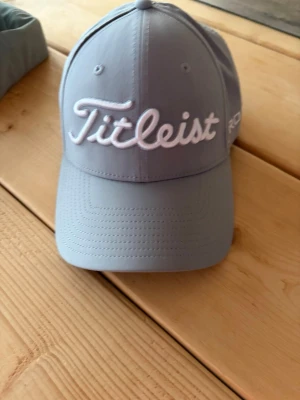 Ljusgrå Titleist keps - Snygg ljusgrå keps från Titleist med broderad vit logga framtill och FJ-detalj på sidan. Klassisk böjd skärm och justerbar passform. Tillverkad i ett lätt och ventilerande material som passar perfekt för golf eller vardagsstil.