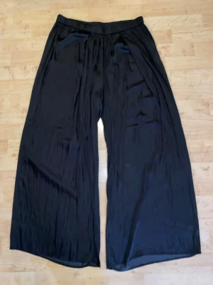 Svarta vida linnebyxor från H&M - Snygga svarta linnebyxor från H&M med bred resår i midjan och riktigt vida ben. Byxorna har en lätt glansig finish och är tillverkade i ett tunt, luftigt material som känns skönt mot huden. Perfekta för en avslappnad och trendig look.