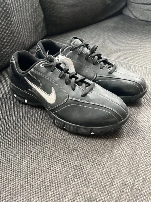 Svarta Nike golfskor med dobbar - Säljer ett par helt oanvända svarta golfskor från Nike med vita detaljer och klassisk swoosh-logga på sidan. Skorna har snörning och slitstarka dobbar på sulan för bra grepp på golfbanan. Materialet är en mix av syntet och textil för komfort och hållbarhet. Lappen sitter kvar på skorna men lådan fås inte med då den är borta tyvärr. Storlek 42,5