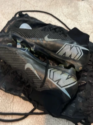 Nike Mercurial Superfly fotbollsskor - Snygga Nike Mercurial Superfly Elite fotbollsskor i svart med silverdetaljer och stor 'M' på hälen. Skorna har strumpliknande skaft och är tillverkade i syntetmaterial för bra passform och grepp. Perfekta för snabba spelare som vill sticka ut på planen. Det är i väldigt bra skick, endast lite smutsiga som enkelt går bort med en tvätt. Nypris Ca 3200 Kvitto finns. Hör av er vid fundering.
