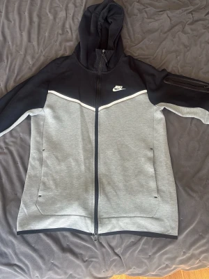 Nike Tech Fleece hoodie svart/grå - Nike Tech Fleece hoodie i svart och grått med hel dragkedja framtill. Stora fickor på sidorna, vit detaljlinje över bröstet och diskret Nike-logga på vänster sida. Huva och dragkedjeförsedd ficka på ärmen. Tillverkad i mjukt och bekvämt material.