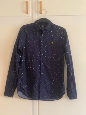 Mörkblå prickig skjorta Lyle & Scott - Mörkblå skjorta från Lyle & Scott med små vita prickar och klassisk krage. Skjortan har långa ärmar, knappar framtill och den ikoniska gula loggan broderad på bröstet. Perfekt för en stilren och avslappnad look.