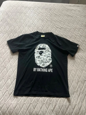 Svart BAPE t-shirt med camotryck - Snygg svart t-shirt från BAPE med det ikoniska apansiktet i grått camomönster och texten 'BY BATHING APE' på bröstet. Klassisk passform och rund hals. Perfekt för dig som gillar streetwear och vill sticka ut. Står storlek L men passar bäst som en M.
