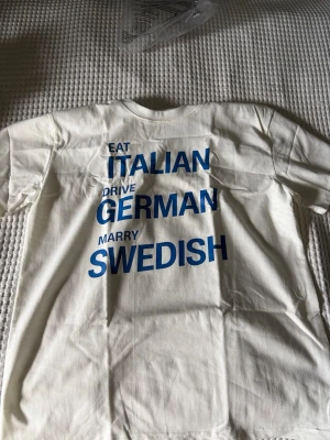 Vit t-shirt med blå text oanvänd - Vit t-shirt i bomull med tryck i blå text: 'EAT ITALIAN, DRIVE GERMAN, MARRY SWEDISH'. Klassisk passform och rund hals. Perfekt för dig som gillar statement-plagg och enkel stil.