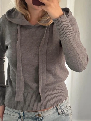 Grå hoodie med ribbade muddar - Mysig grå hoodie med huva och långa snören. Tröjan har ribbade muddar vid ärmslut och nederkant, vilket ger en snygg och avslappnad look. Tillverkad i mjukt stickat material som känns skönt mot huden. I mycket bra skick, storlek M men passar som S.